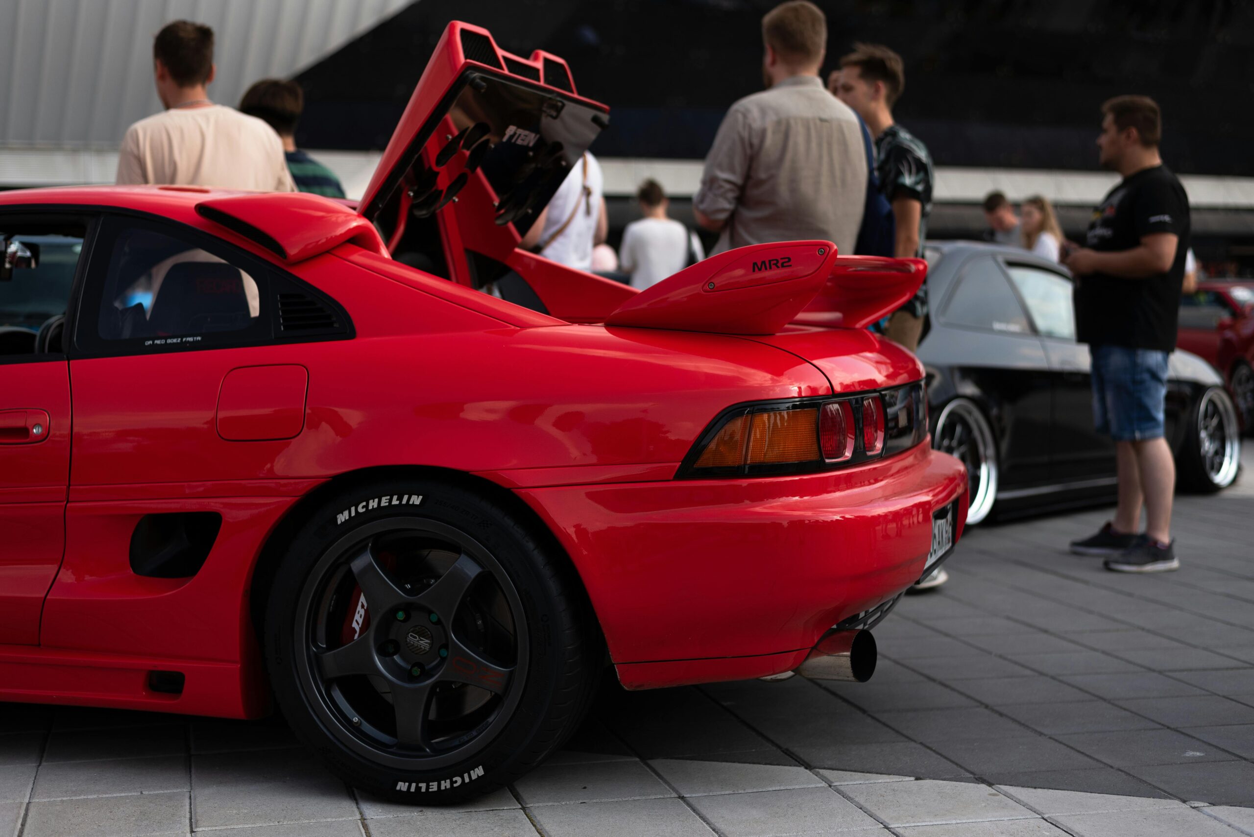 Red Toyota MR2 showcased at an urban car event with people in background.